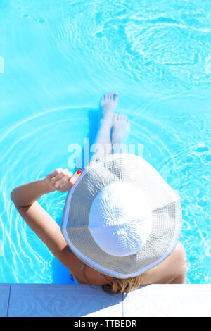 unrecognizable woman with hat relaxing by the swimming pool in a luxury ...