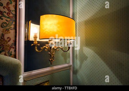 Warm hotel wall lamp reflected in a mirror, casting soft light and shadow on patterned wallpaper in an elegant interior. Stock Photo
