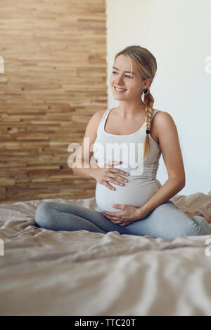 Happy young pregnant woman bonding with her unborn child sitting on a bed cradling her baby bump with her hands looking sideways with a tender smile o Stock Photo