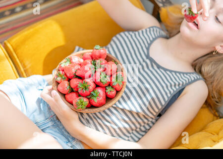 Attractive young woman laying on sofa in a home family room living room, eating fresh strawberry. Girl eating healthy food at home, interior. Well bei Stock Photo