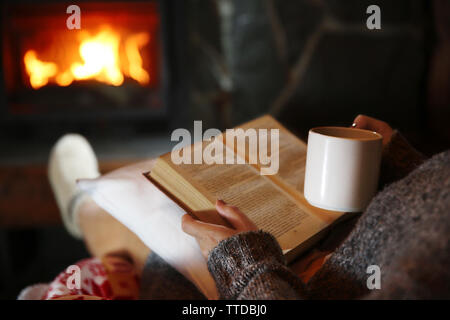 Woman resting with cup of hot drink and book near fireplace Stock Photo