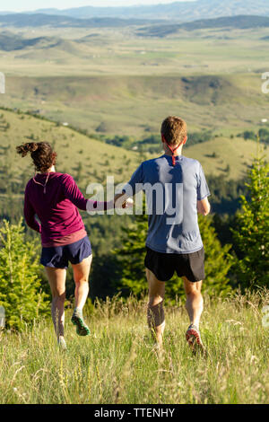 Happy millennial couple holding hands and frolicking down a grass covered hill in the early morning light. Rocky Mountains in June. Montana, USA. Stock Photo