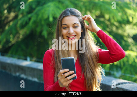 Surprised young woman using smart phone outdoors. Excited urban girl using new mobile phone app in city park. Stock Photo