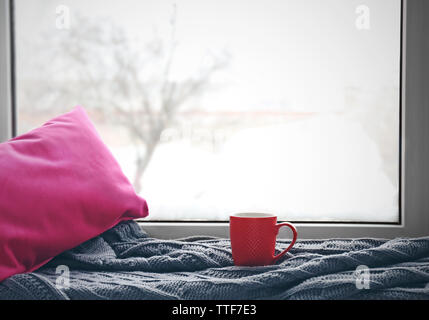 Red cup of tea, warm knitted plaid and pink pillow on windowsill, close up Stock Photo