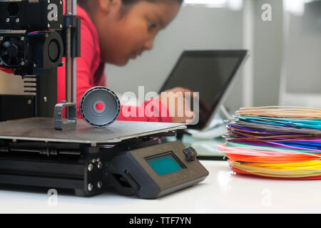 A 3d printer with two 3d models that have finished printing on the heatbed, colorful filament and a young girl in the background using a tablet. Stock Photo