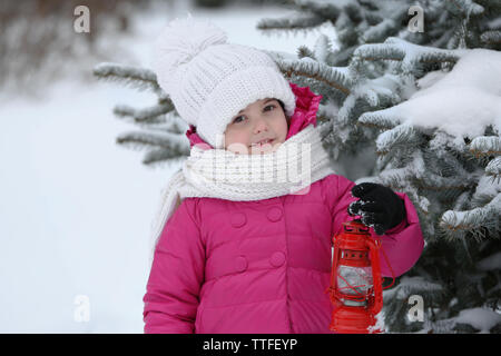 Little girl with winter clothes holding red lantern near fir tree in snowy park outdoor Stock Photo