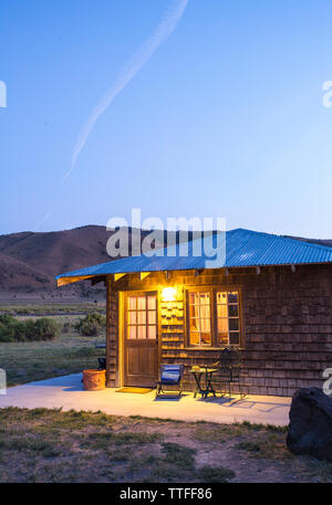 A small cabin with exterior light glowing under a deep blue dusk sky w Stock Photo