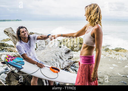Happy male surfer holding woman's hand while sitting on driftwood at beach Stock Photo