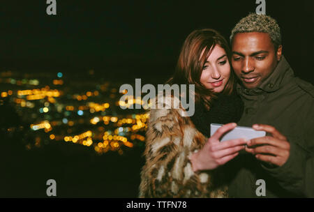 Happy young couple looking at smart phone while standing against cityscape Stock Photo