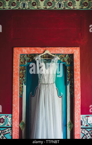 Beautiful white dress on hanger on traditional balinese textured red wooden door. Wedding concept. Stock Photo