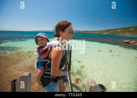 Happy mother carrying son in carriage while standing on pier at beach against clear blue sky Stock Photo