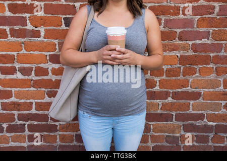 Midsection of pregnant woman holding coffee cup while standing against brick wall Stock Photo