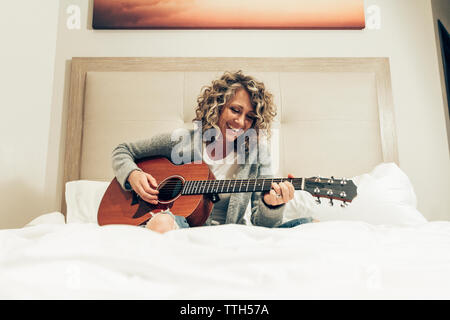 Smiling businesswoman playing guitar while sitting on bed in hotel room Stock Photo