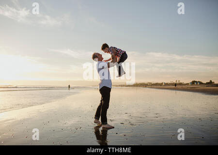 Playful father lifting son while playing at beach against sky during sunset Stock Photo