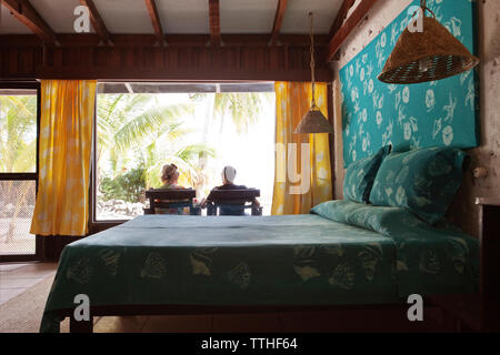 Rear view of couple relaxing on lounge chairs seen through bedroom window Stock Photo