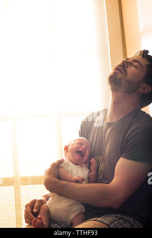 Frustrated father holding crying baby daughter against window at home Stock Photo