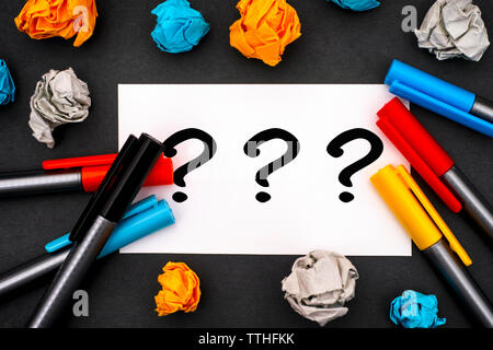 Three question marks on white paper blank with colour pens and crumpled paper. Black background. Stock Photo