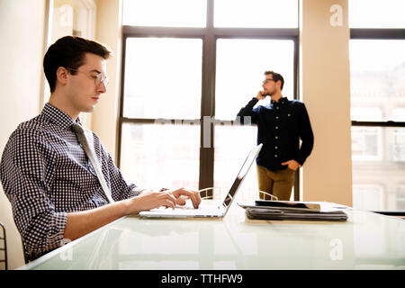 businessman using laptop at table with colleague using phone in background Stock Photo