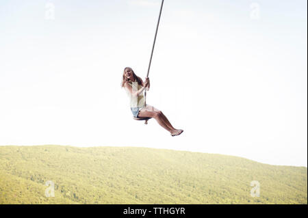 Low angle view of cheerful woman swinging on rope against clear sky Stock Photo