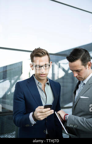 businessman using phone while colleague checking time on watch while standing outdoors Stock Photo