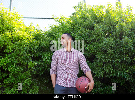 Happy young man holding basketball while standing against plants Stock Photo
