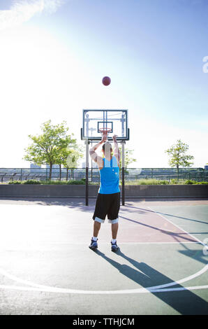 Rear view of man throwing basketball in hoop against sky during sunny day Stock Photo