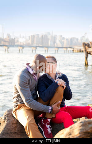 Boyfriends kissing while sitting on rocks against river Stock Photo
