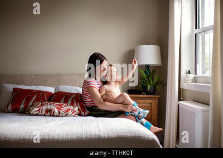 Mother dressing playful son while sitting on bed Stock Photo