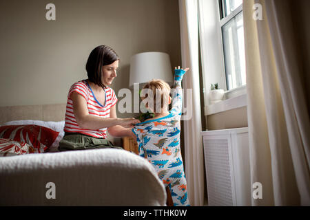 Mother dressing son while sitting on bed in room Stock Photo