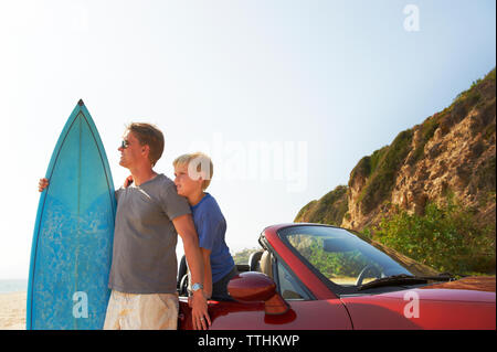 Father and son with surfboard by car against sky Stock Photo