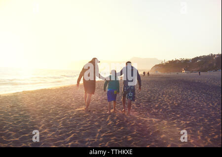 Rear view of family walking at beach against clear sky during sunset Stock Photo