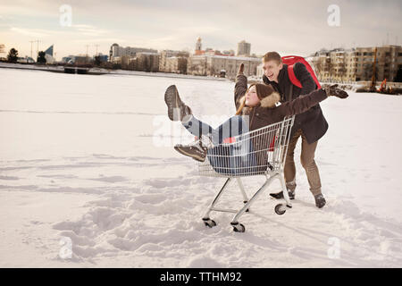 Happy man pushing girlfriend sitting in shopping cart on snow covered field Stock Photo