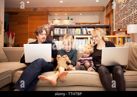Siblings using mobile phones while sitting with parents on sofa at home Stock Photo