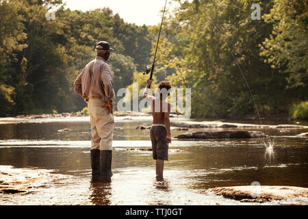 Rear view of boy fishing by grandfather standing by lake Stock Photo