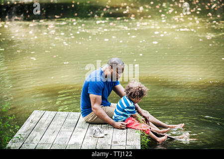 Father and son sitting on jetty with feet in lake Stock Photo