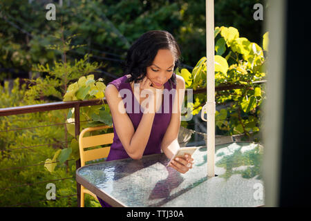 Woman using smart phone while sitting on chair by table in balcony Stock Photo