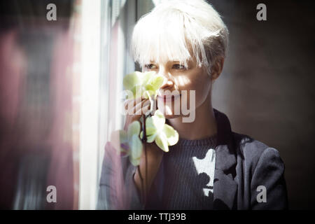 Thoughtful woman holding flowers while looking through window at home Stock Photo
