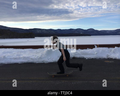 Full length side view of boy skateboarding on road by river during winter Stock Photo
