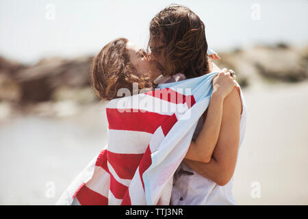 Side view of woman wrapped in blanket while kissing man on beach Stock Photo