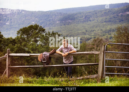 Rancher with saddle leaning on railing while standing on grassy field against mountain Stock Photo