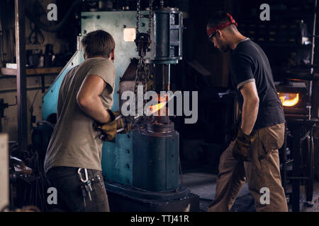 Blacksmith standing by coworker forging rod in machinery at workshop Stock Photo