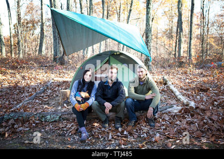 Portrait of friends sitting on fallen tree trunk by tent in forest Stock Photo
