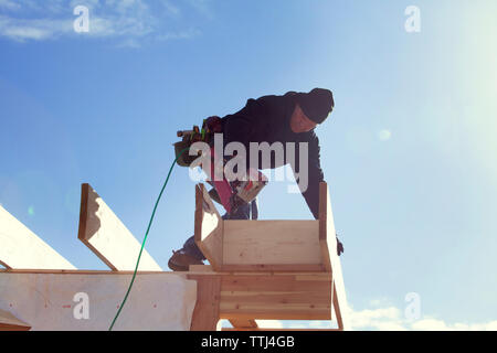 Low angle view of worker with drill standing on roof beam against sky Stock Photo