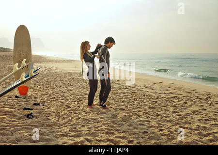 Woman assisting man in wearing wetsuit while standing at beach Stock Photo