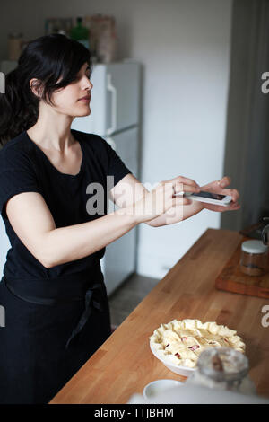 Woman photographing pie through mobile phone while standing in kitchen Stock Photo
