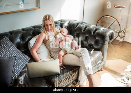 Mother using laptop computer while sitting with baby boy on sofa at home Stock Photo