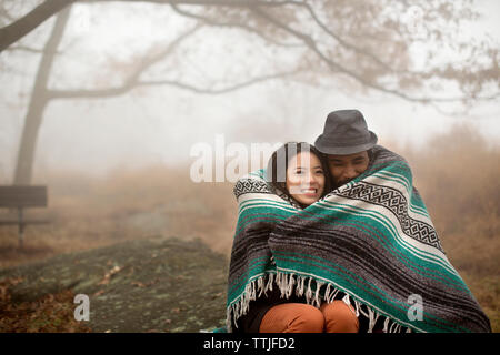 Happy couple wrapped in blanket while sitting on rock in forest Stock Photo