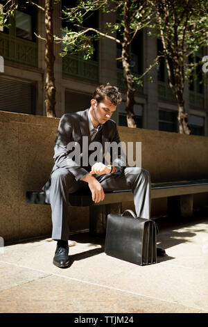 Man with briefcase looking at wristwatch while sitting on bench against building Stock Photo
