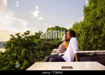 Side view of couple sitting on retaining wall against sky Stock Photo