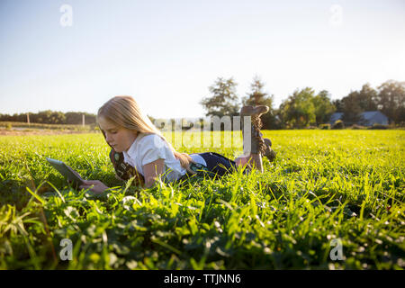 schoolgirl using tablet computer while lying on grassy field against clear sky Stock Photo